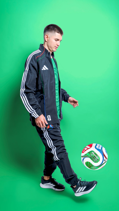 A young man in a sports jacket and pants is juggling a soccer ball against a vibrant green background.