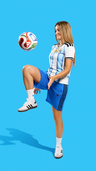 A young woman in a sports uniform is kicking a soccer ball against a bright blue background.