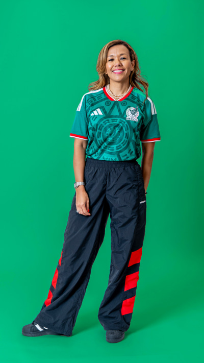 A smiling person wearing a green and white sports jersey and navy blue pants stands in front of a vibrant green background.