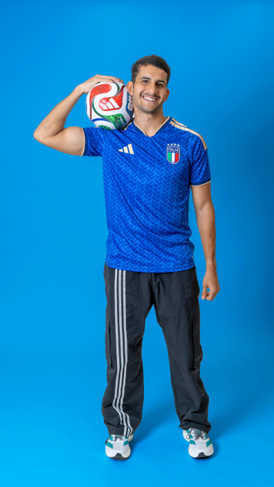 A smiling man wearing an Italian national team soccer jersey and holding a soccer ball stands in front of a vibrant blue background.