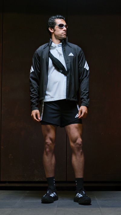 A man wearing a black jacket, white shirt, and black shorts stands in a dark setting, his muscular physique and confident posture commanding attention.