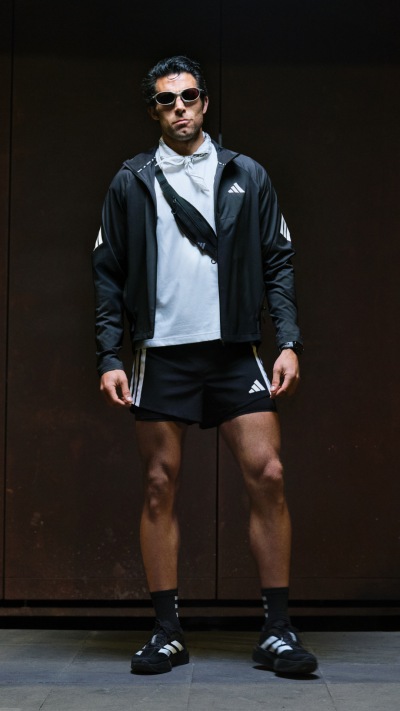 A person wearing a black jacket, white shirt, and black shorts stands in front of a dark background.