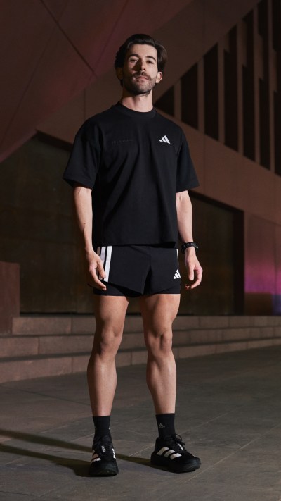 A muscular man wearing a black athletic outfit stands in a dimly lit indoor setting, with a plain background behind him.