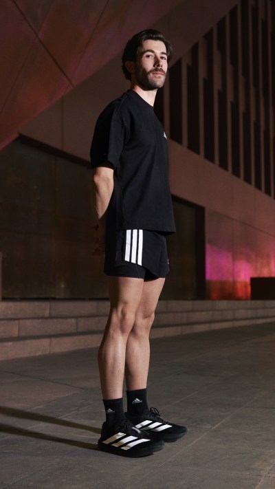 A person with a beard wearing a black shirt and shorts stands in a dimly lit area with a patterned wall in the background.