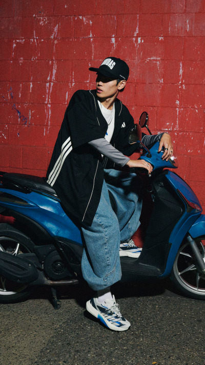 A person wearing a black jacket, cap, and jeans is sitting on a blue motorcycle in front of a red brick wall.