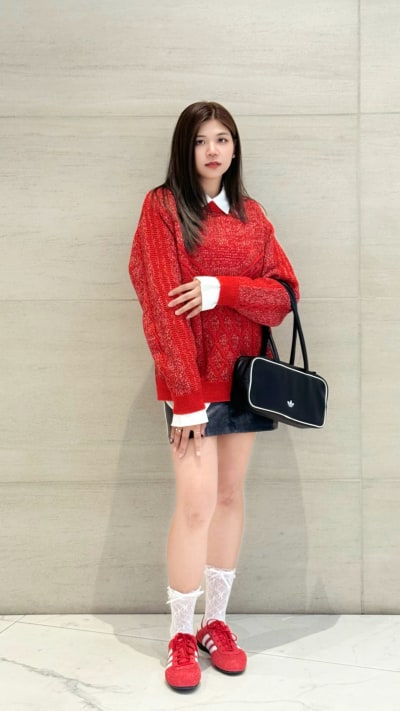 A young woman wearing a bright red sweater, white socks, and red sneakers stands in front of a plain white wall, holding a black handbag.