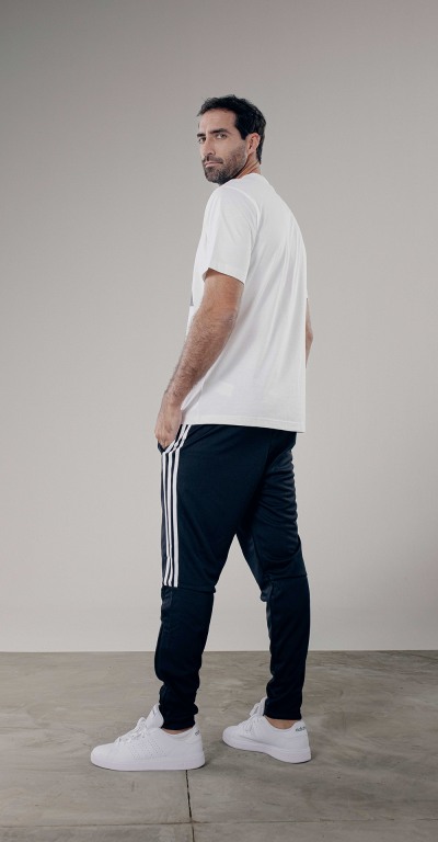 A man wearing a white t-shirt, black pants, and white sneakers stands in a plain, light-colored setting.
