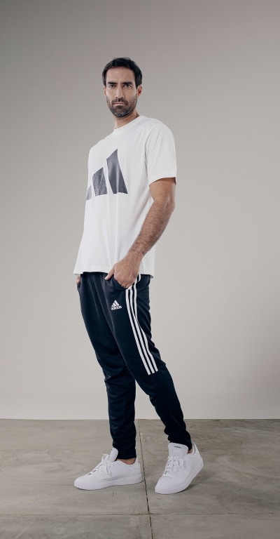 A man wearing a white t-shirt with a graphic design, black Adidas sweatpants, and white sneakers stands in a plain, gray-toned setting.