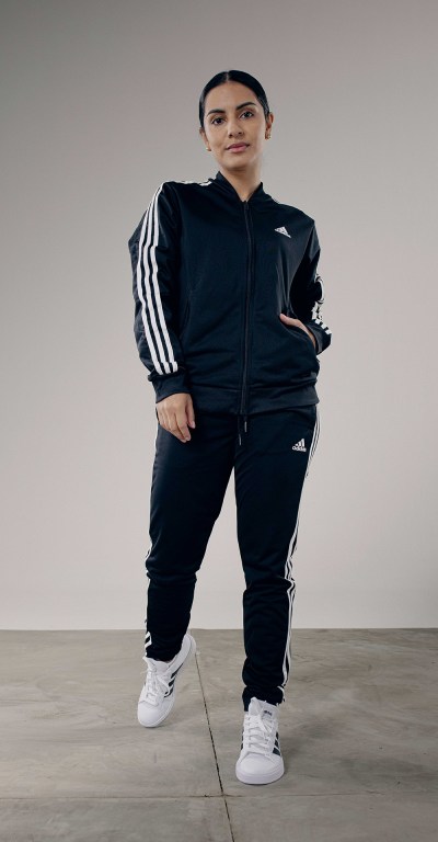 A person wearing a black tracksuit with white stripes stands in a plain, gray-toned setting.