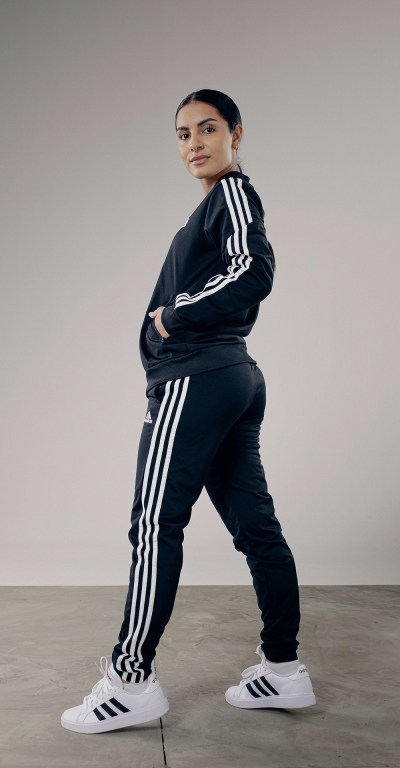 A person wearing a black tracksuit with white stripes stands in a plain, light-colored setting.