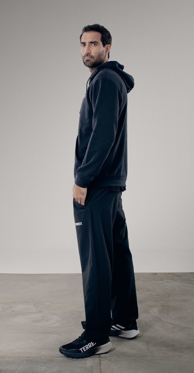 A man wearing a black hooded sweatshirt and pants stands in a plain, gray-toned setting.