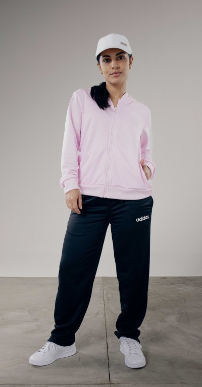 A person wearing a pink hooded sweatshirt and black Adidas sweatpants stands in a plain, gray-toned setting.