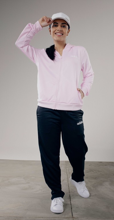 A smiling person wearing a pink jacket, black pants, and white sneakers stands in a plain, light-colored setting.