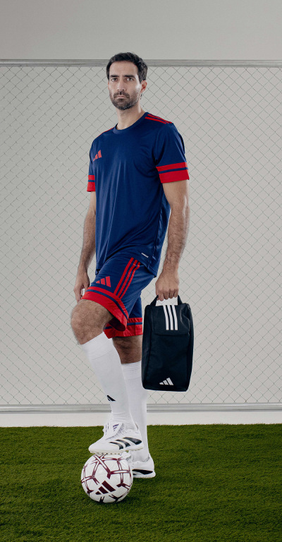 A man in a navy blue and red sports jersey stands on a grassy field, holding a soccer ball and a sports bag.