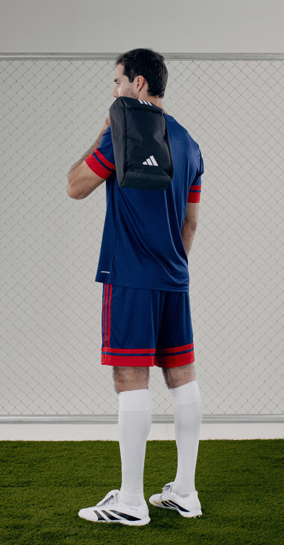 A person wearing a blue sports jersey and shorts, carrying a backpack, stands on a grassy field with a white wall in the background.
