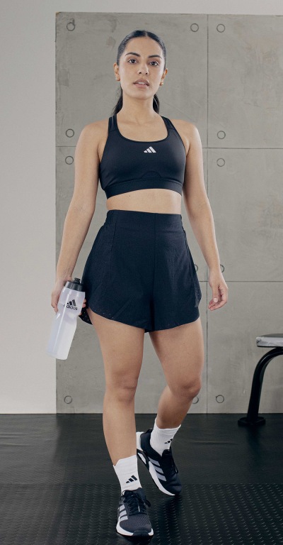 A woman with a curvy figure wearing a black sports bra and shorts stands in a gym setting with a gray concrete wall in the background.