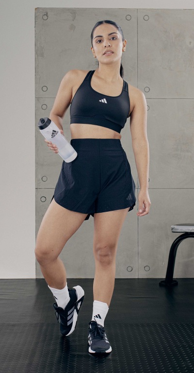 The image shows a woman wearing a black sports bra and shorts, standing in a fitness studio with a gray background.