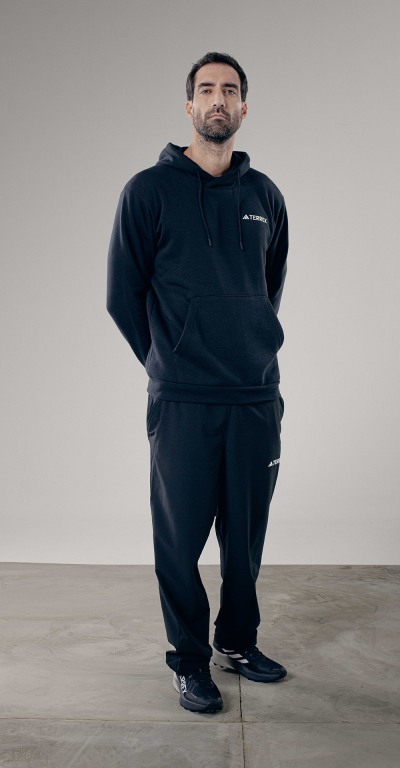 A man wearing a black hooded sweatshirt and pants stands on a plain gray background.