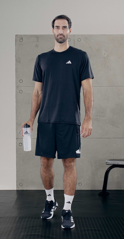 A man wearing a black t-shirt, black shorts, and black and white sneakers stands in a room with concrete walls and floors.