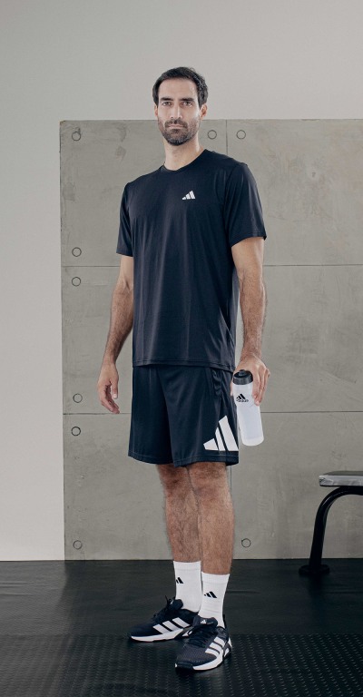 A man wearing a black t-shirt and shorts stands in a room with a gray background, holding a water bottle.