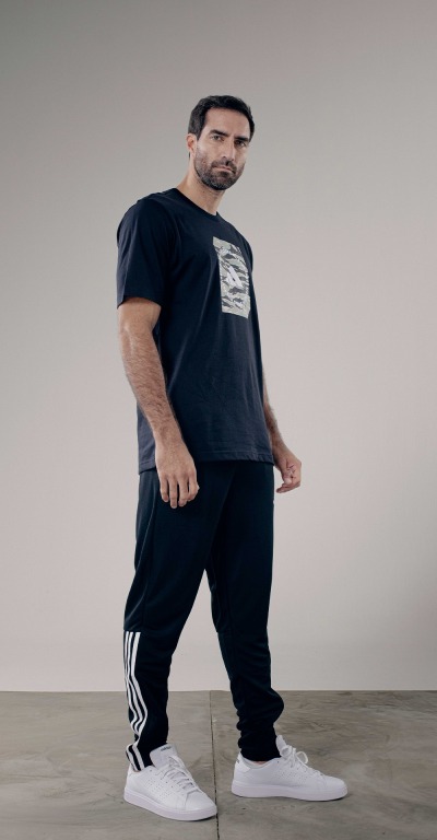 A man wearing a black t-shirt and black pants with white sneakers stands in a plain, light-colored setting.