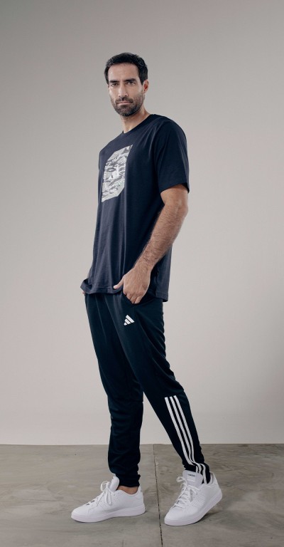 A man wearing a black t-shirt, black pants, and white sneakers stands in a plain, light-colored setting.