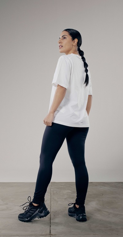 A woman wearing a white t-shirt and black leggings stands on a gray floor against a plain background.