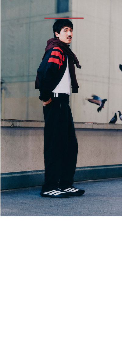 A person wearing a black robe and red-striped uniform stands in a room with a glass wall, with birds visible in the background.
