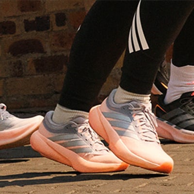 The image shows a pair of white and orange sneakers with three stripes on the side, standing on a brick wall background.