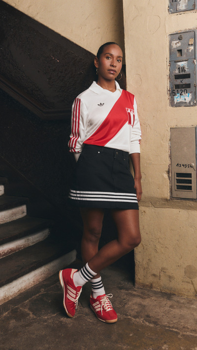 A young person wearing a red and white sports jersey, black shorts, and red sneakers stands on a set of stairs in front of a weathered wall.