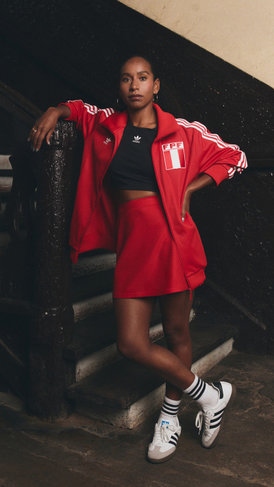A young person wearing a red Adidas jacket and shorts, standing on a staircase against a dark background.