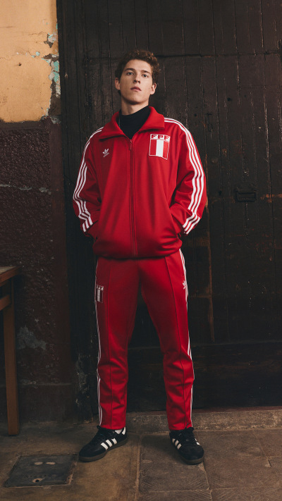 A young man wearing a red tracksuit stands in front of a weathered, wooden wall.