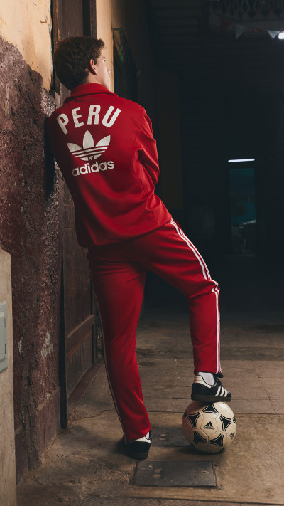 A person wearing a red Adidas tracksuit is standing in a dimly lit alley, with a soccer ball at their feet.