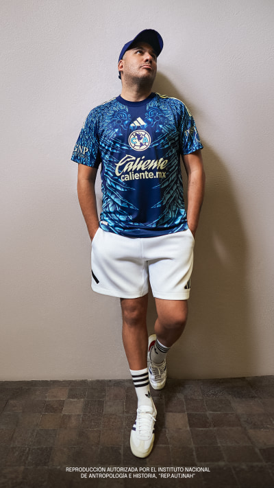 A person wearing a blue and white patterned sports jersey and white shorts, standing against a plain wall.