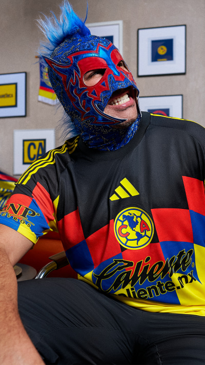 A colorful and vibrant wrestling mask with intricate designs is worn by a person in a black and red jersey, with various framed images visible in the background.