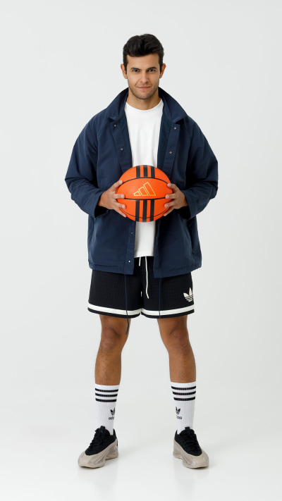 A young man wearing a navy blue jacket, white t-shirt, black shorts, and white socks with black shoes, holding an orange American football in front of a plain white background.