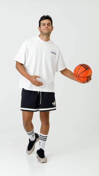 A young man wearing a white t-shirt, black shorts, and sneakers stands holding a basketball in a plain white background.