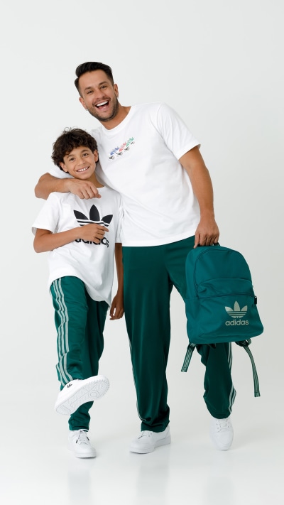 Two individuals, a man and a child, are wearing matching Adidas clothing and smiling at the camera. The background is a plain white surface.