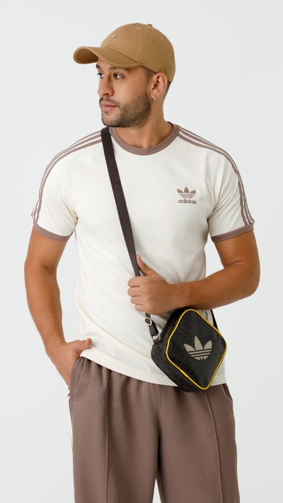 A man wearing a white t-shirt, brown pants, and a baseball cap stands in the foreground, holding a small black and yellow bag.