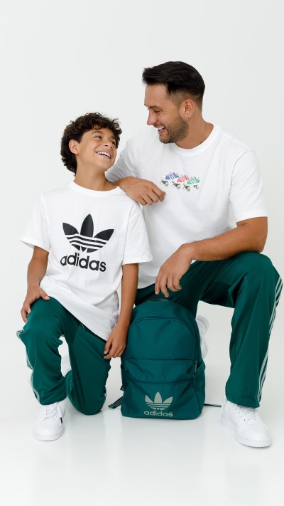 A smiling young boy and a man wearing Adidas clothing are sitting together on the floor, surrounded by Adidas backpacks.