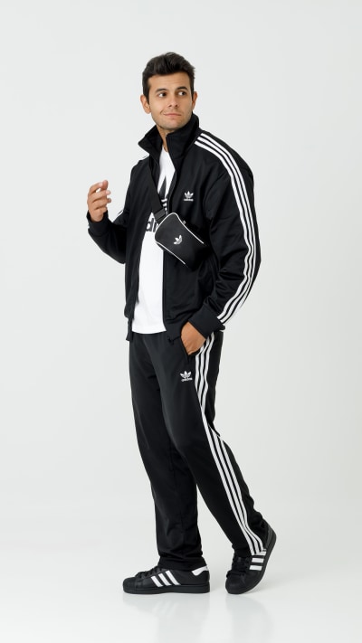 A young man wearing a black and white Adidas tracksuit stands against a plain white background, with a confident and casual posture.