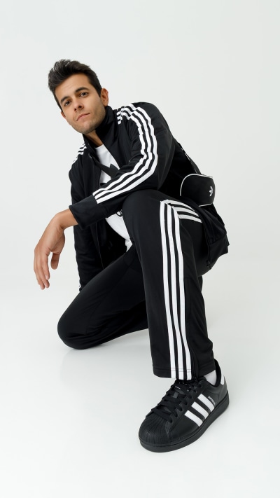 A young man wearing a black tracksuit with white stripes sits on the floor against a plain white background.