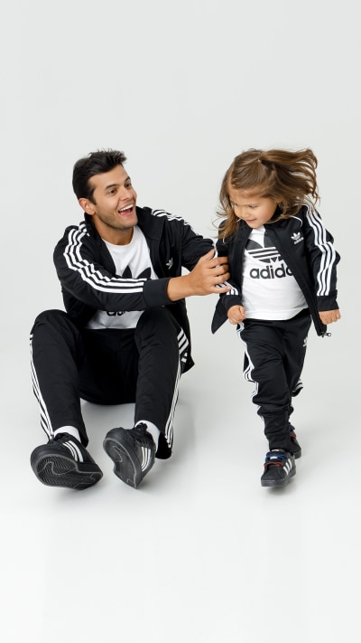 A man and a young girl, both wearing Adidas sportswear, sit together on a plain white background, the man smiling and interacting with the girl.