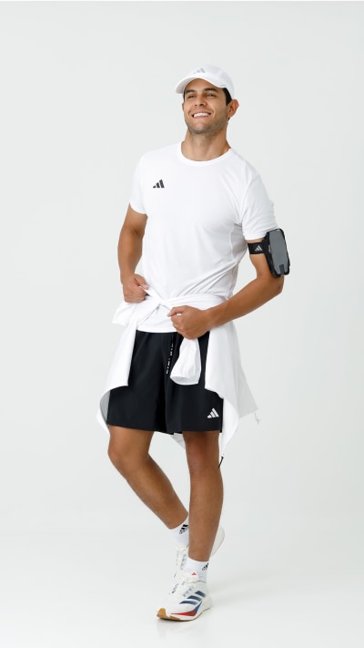 A smiling man wearing a white t-shirt, black shorts, and white sneakers stands against a plain white background.