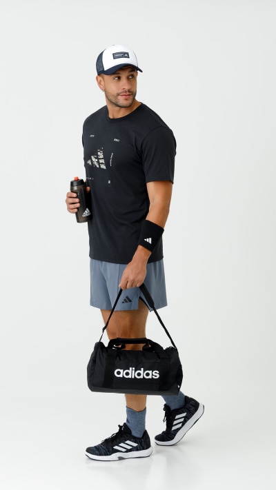A person wearing a black t-shirt, gray shorts, and a white and black baseball cap, holding a black sports bag and standing against a plain white background.