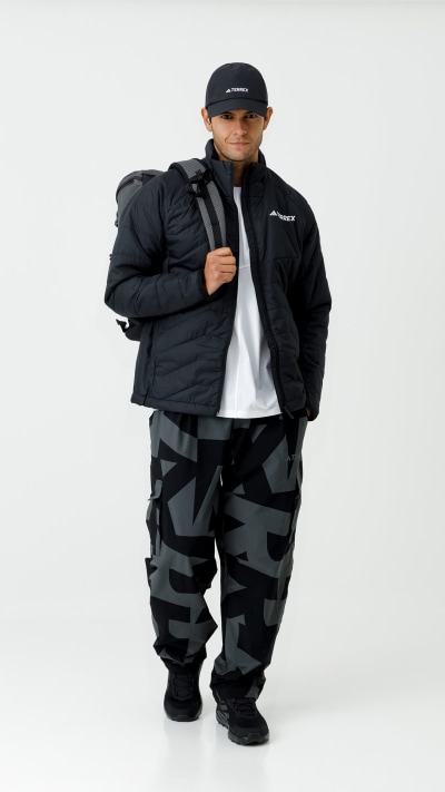 A person wearing a black jacket, hat, and camouflage pants stands against a plain white background.
