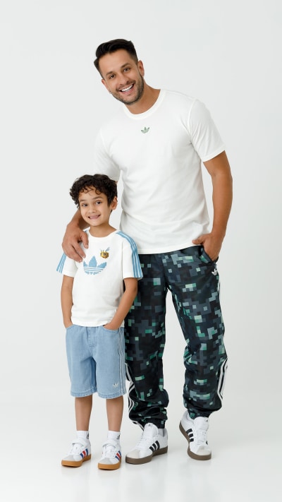 A smiling man in a white t-shirt stands next to a young boy wearing a similar t-shirt, both wearing casual clothing and posing against a plain white background.