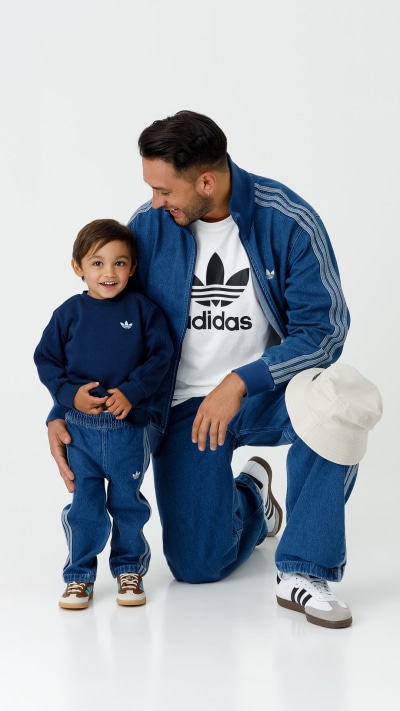 A man wearing an Adidas tracksuit is sitting on the floor and embracing a young child also wearing an Adidas outfit, both smiling and appearing to have a warm, affectionate interaction.