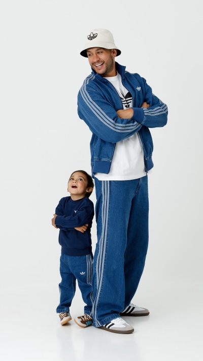 A smiling man wearing a white hat and a blue tracksuit stands next to a young child also wearing a blue tracksuit, both appearing happy and relaxed against a plain white background.