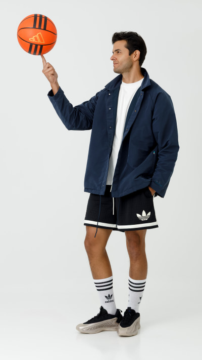 A man in a navy blue jacket and shorts is holding an orange rugby ball, standing against a plain white background.